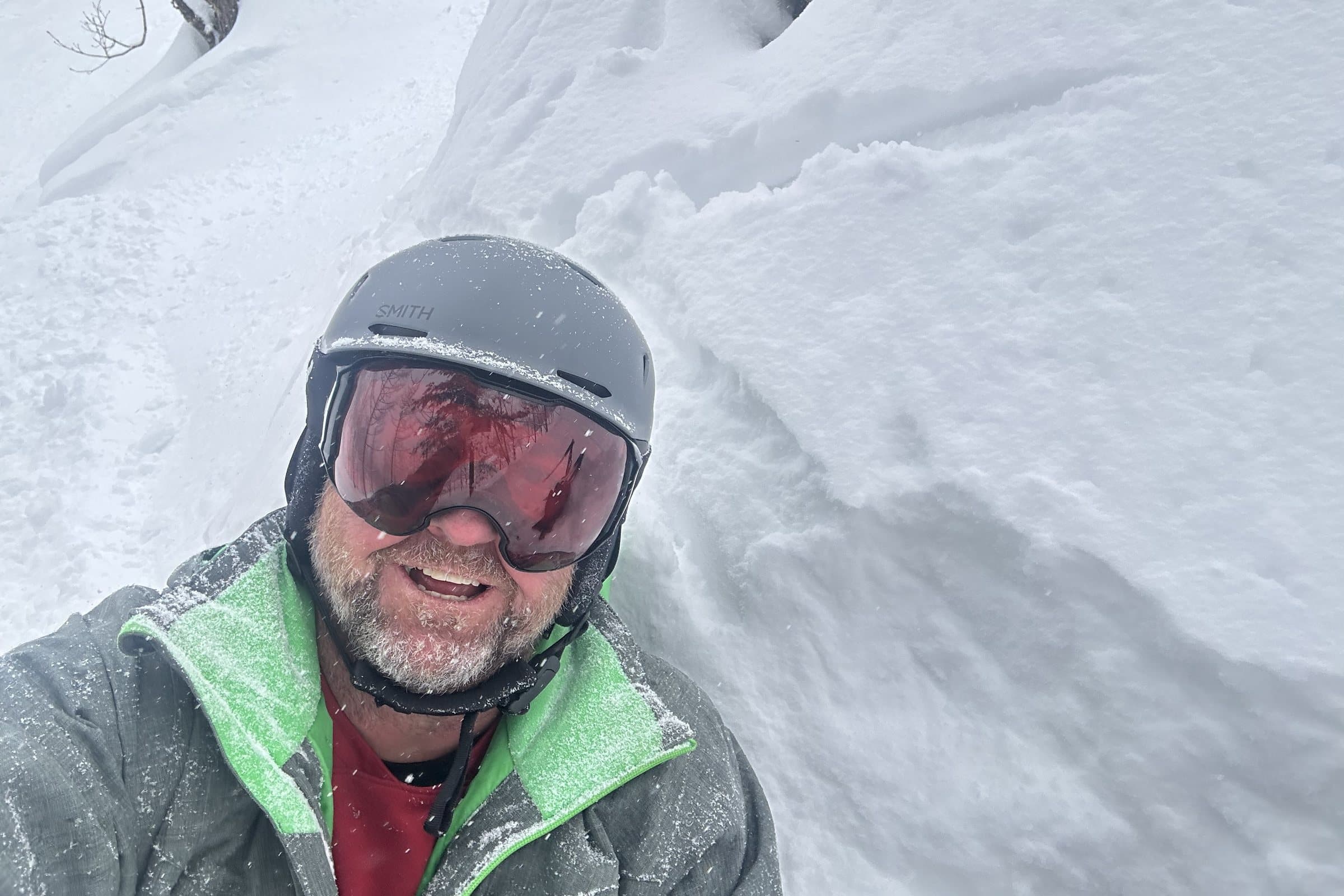 Angus Gordon in deep untracked snow among trees, helmet and goggles on, grinning, mid-run off-piste.
